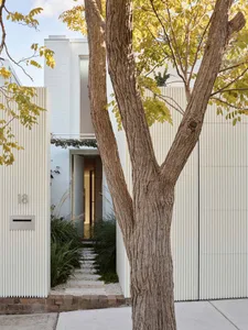 A modern entryway featuring a white-paneled wall, brick pathway, and a tree with yellow leaves.