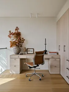 Modern workspace featuring a wooden desk and chair with a vase of autumn leaves on the desk.