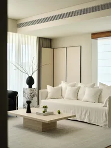 A minimalist residential room featuring soft, neutral-colored furniture and accents, with large windows allowing natural ligh