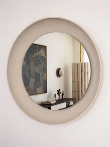 A modern circle wall mirror with a textured concrete frame hanging on a light-colored wall, reflecting a living room with a w