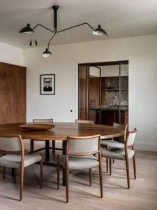 A modern dining room with a round walnut table surrounded by upholstered chairs, set against a white wall and adjacent to a k