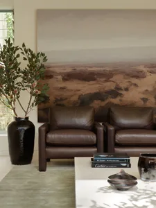 A modern living room with brown leather chairs, a green plant in a black vase, a large abstract artwork, and a coffee table.