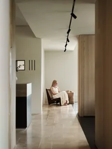 A woman in a white dress sits casually on a wooden chair in a modern residential space. Minimalist design with exposed concre