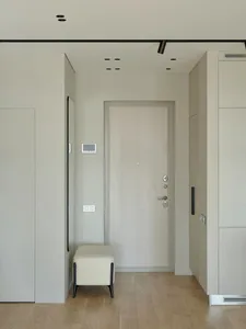 A minimalist entrance area featuring a light-colored wooden door, a beige upholstered bench, and a sleek black light fixture.