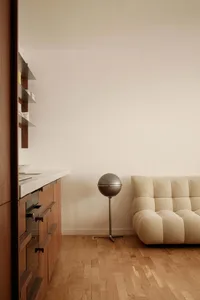 A minimalist living room featuring a beige tufted sofa, a spherical floor lamp, and wooden cabinets against a plain wall on a