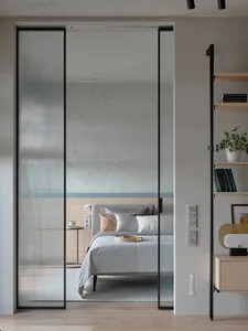 A modern bedroom featuring a clear glass wall, wooden floor, white bed with grey bedding, and a floating shelf with books and