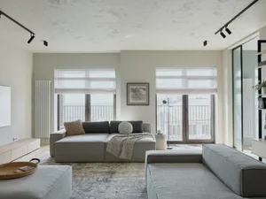 A modern living room featuring neutral-toned sofas under track lighting and large windows with white roman shades.