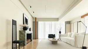 A modern minimalist living room featuring a marble fireplace, white sofa, white curtains, and a large black armchair.