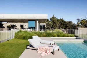 A modern residential exterior featuring a concrete patio area with two white lounge chairs and a swimming pool surrounded by 