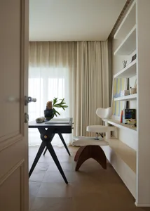 A modern home office space featuring a wooden table and a white desk chair, with a built-in bookshelf and a large window with