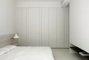 Modern minimalist bedroom with white furniture, concrete walls, and a single light source.