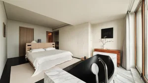 A minimalist bedroom featuring a large bed with a wooden bed frame, marble flooring, and a glass-enclosed bathtub.