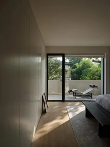 A minimalist bedroom with a bed, black bench, and a view of a balcony with a concrete wall and greenery.