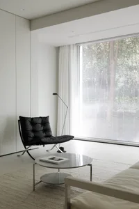 Modern minimalist living room with a black leather chair, white coffee table, and large window with blinds.
