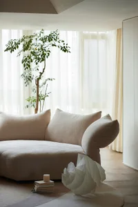 A minimalist living room featuring a boucle sofa, a potted plant, and a decorative paper swan on a rug.