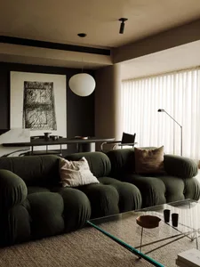 A modern, dark living room with a green fabric sofa, a glass coffee table, and a large window with white curtains.