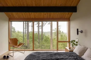 A minimalist bedroom with a large window overlooking a forest, featuring a bed with a grey blanket and a light brown leather