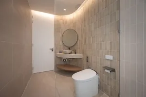 A modern bathroom interior with minimalist design, featuring beige tiles, a round mirror, a sink, a toilet, and a wooden shel
