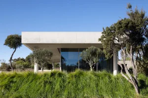 A modern residence with a white concrete exterior, large glass windows, and surrounded by tall grass and trees.