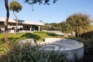 A modern home with a concrete outdoor space featuring a circular seating area, greenery, and a building with large glass wind