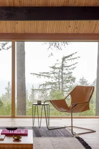 Modern living room with a wooden floor, large windows, and a comfortable chair facing a scenic landscape.