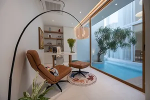 Modern living room with a glass wall overlooking a poolside area, featuring a brown leather chair, ottoman, and circular rug 