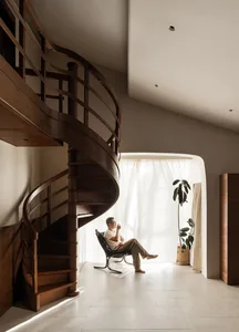 A modern living room with a wooden spiral staircase and a person sitting on a chair by a window.