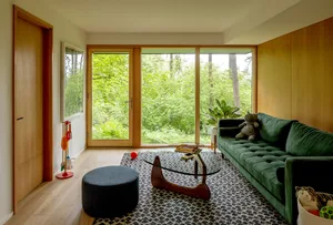 A modern minimalist living room featuring a green sofa, glass coffee table, and wooden flooring with a view of greenery outsi