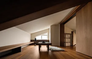 A minimalist living space with a wood-finished floor, featuring a bench, a table with a lamp, and a rug.