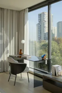 A modern office space featuring a transparent glass desk and chair, with minimalist decor and a view of tall buildings and gr