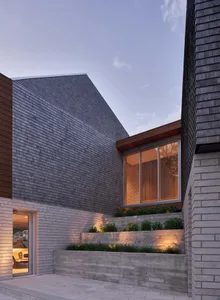 Modern residential exterior featuring wood shingles, concrete walls, and large windows with illuminated interior.