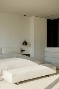 A minimalist bedroom with white furniture and a large window.