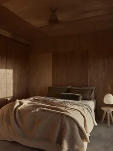 A minimalist bedroom featuring wooden walls, a bed with cream-colored bedding, and a small wooden side table with a lamp.