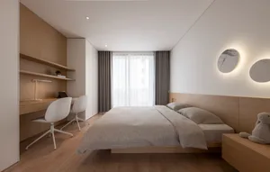 Minimalist bedroom with a wooden bed, fabric bedding, and a white chair with a desk lamp.