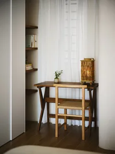 A minimalist home office with a wooden desk, chair, and a lantern lamp, featuring sheer curtains and a bookshelf.