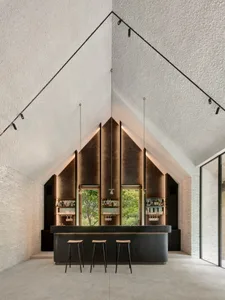 A modern bar interior featuring a concrete and brass counter with three stools, set against a textured wall and glass wall.
