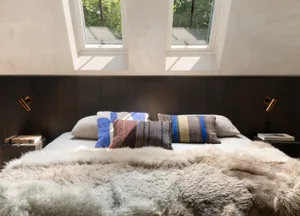 A cozy modern bedroom with textured pillows and blankets, window light streaming in, and warm lighting.