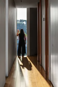 A person standing in a hallway, looking out through a window with a view of trees and sunlight streaming in through the floor