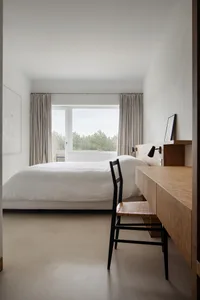 A minimalist bedroom featuring a wooden bed frame and matching desk, with light curtains and a view of trees through the wind