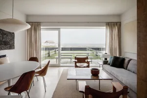 A modern, minimalist living room features glass windows overlooking the ocean, with a wooden table, leather chairs, and a gre