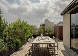 A modern outdoor terrace featuring a concrete dining table and wooden chairs surrounded by lush green plants.