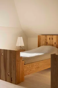 A minimalist bedroom featuring a wooden bed frame with geometric cutouts, a matching wooden nightstand, and a white lamp with