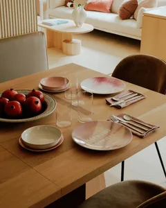A modern minimalist dining room setup with wooden furniture, bowls, glasses, pink plates, cutlery, and pomegranates.