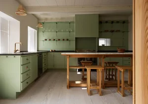 An elegant, modern residential kitchen featuring green painted walls, wooden stools, a marble countertop, and open shelves.