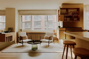 A modern living room with a glass coffee table, two white chairs with wooden legs, a wooden floor, and a large window overloo