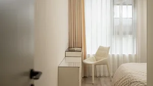 A minimalist bedroom with white furniture, featuring a bed with striped bedding and light-colored curtains.