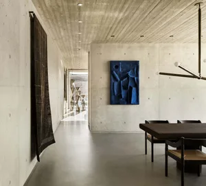 A modern interior space featuring a concrete wall, a blue abstract sculpture, and a wooden dining table with chairs.