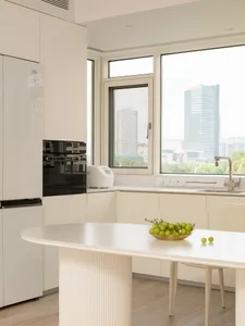 A modern minimalist kitchen featuring white laminate cabinets, a sleek countertop, and a view through a large window.