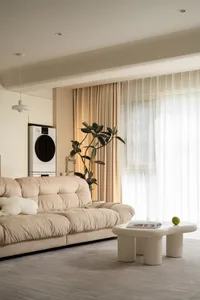A minimalist living room with a beige sofa, a white coffee table with a green apple on it, and a large window with sheer curt