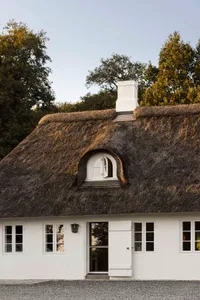 A white house with a thatched roof and a chimney, set against a backdrop of trees and clear sky.